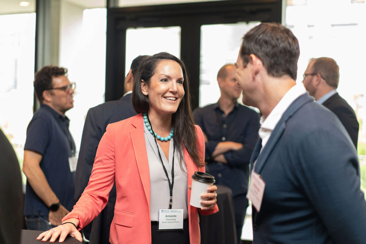 A small group of people engaging in conversation at a networking event. One person in a coral blazer and a conference badge is holding a coffee cup, smiling while talking to another attendee.