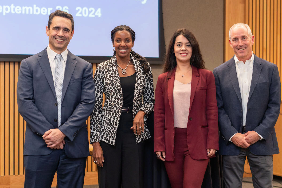 A group of four people standing in business attire, smiling at the camera, with a presentation screen displaying the date September 26, 2024, in the background.