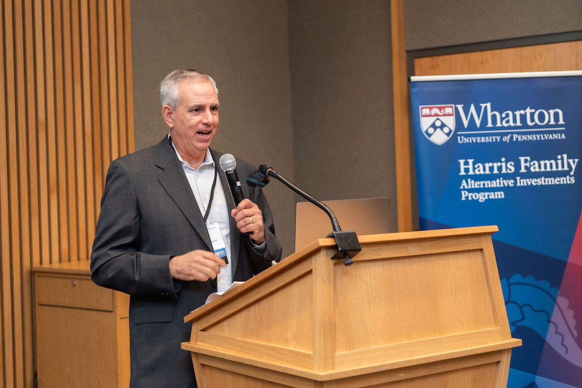 A person in a suit is speaking into a microphone at a podium. A banner for the Wharton University of Pennsylvania Harris Family Alternative Investments Program is in the background.