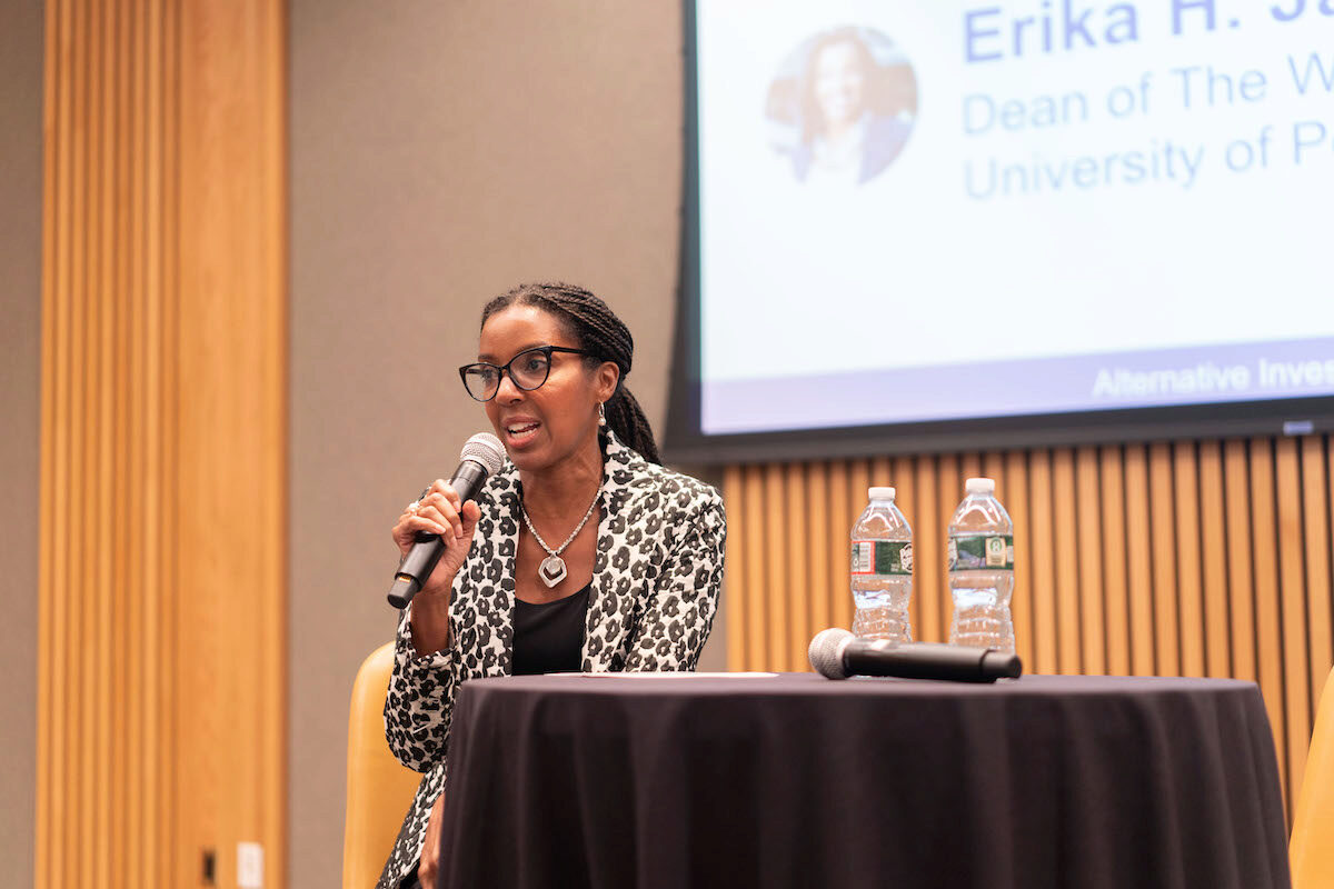 A person speaking into a microphone during a presentation or panel discussion. They are sitting at a table with bottled water and another microphone. A partial presentation slide is visible in the background.