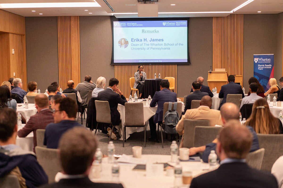 The image shows a conference or seminar setting with several attendees seated at round tables facing a speaker. The speaker is at the front of the room with a presentation screen displaying information about Erika H. James, Dean of The Wharton School of the University of