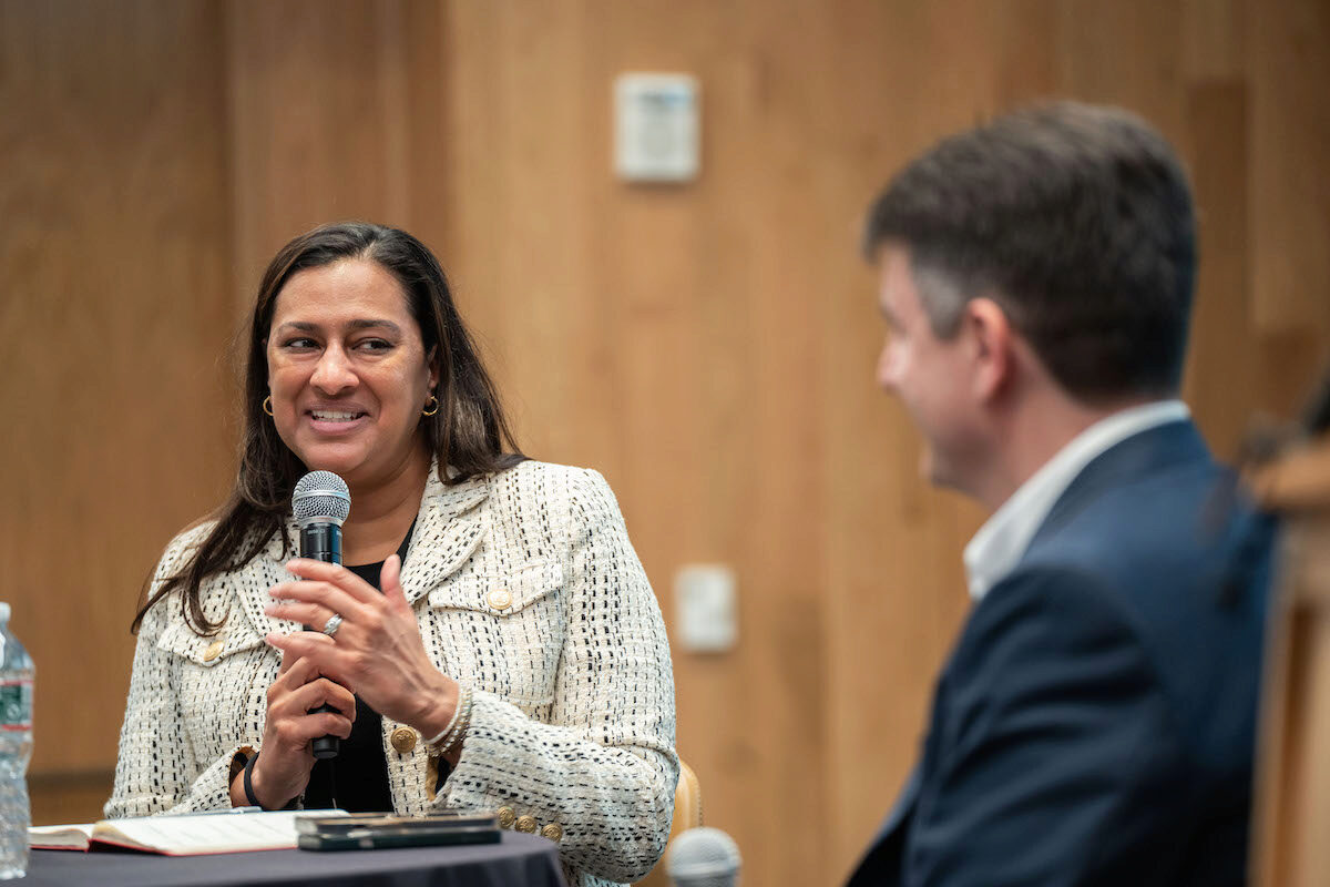 A person holding a microphone is speaking to another person who is listening and smiling. They are in a professional setting, possibly at a conference or meeting.