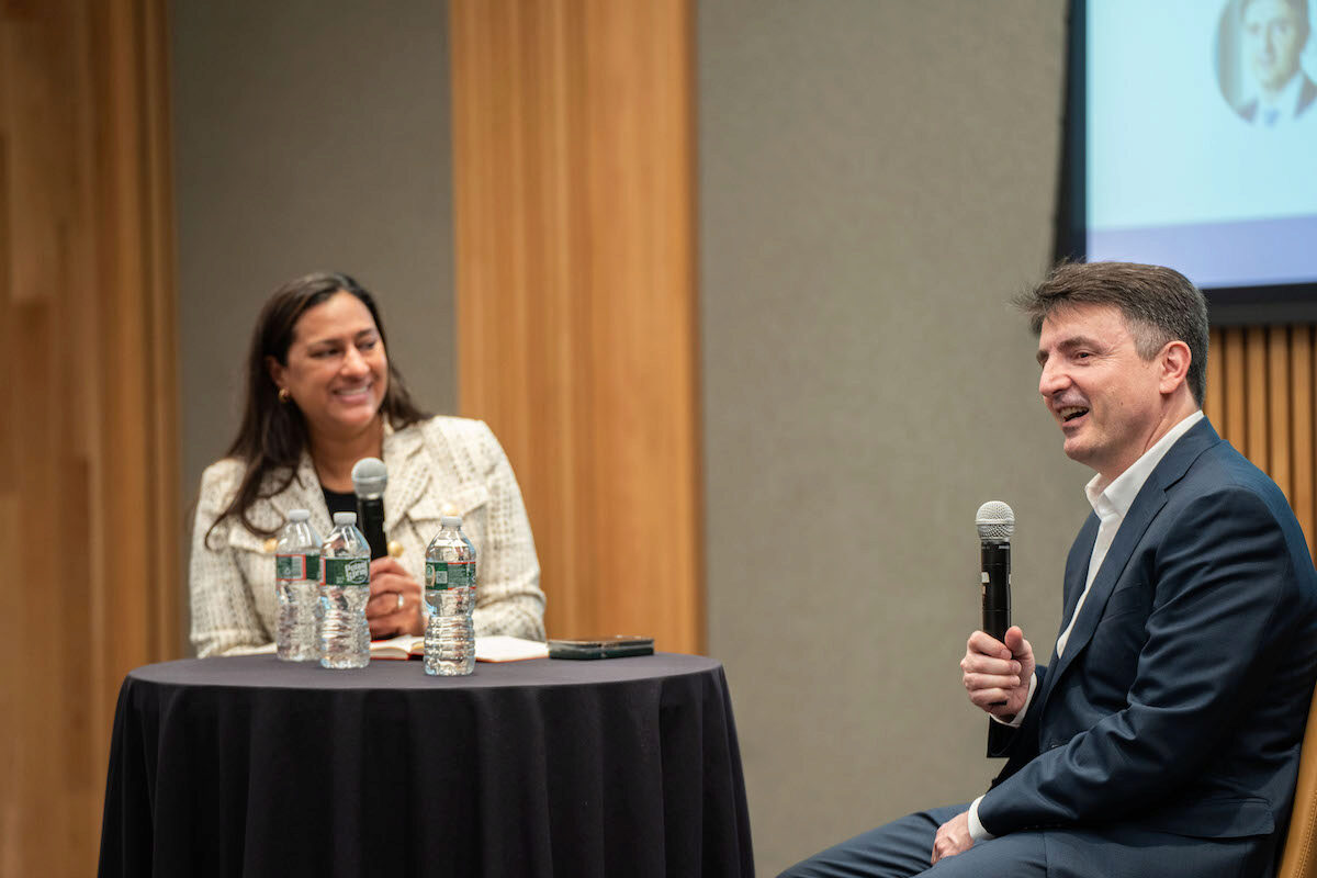 Two people are sitting and talking in a professional setting with microphones. There are water bottles on a table between them, suggesting a discussion or interview.