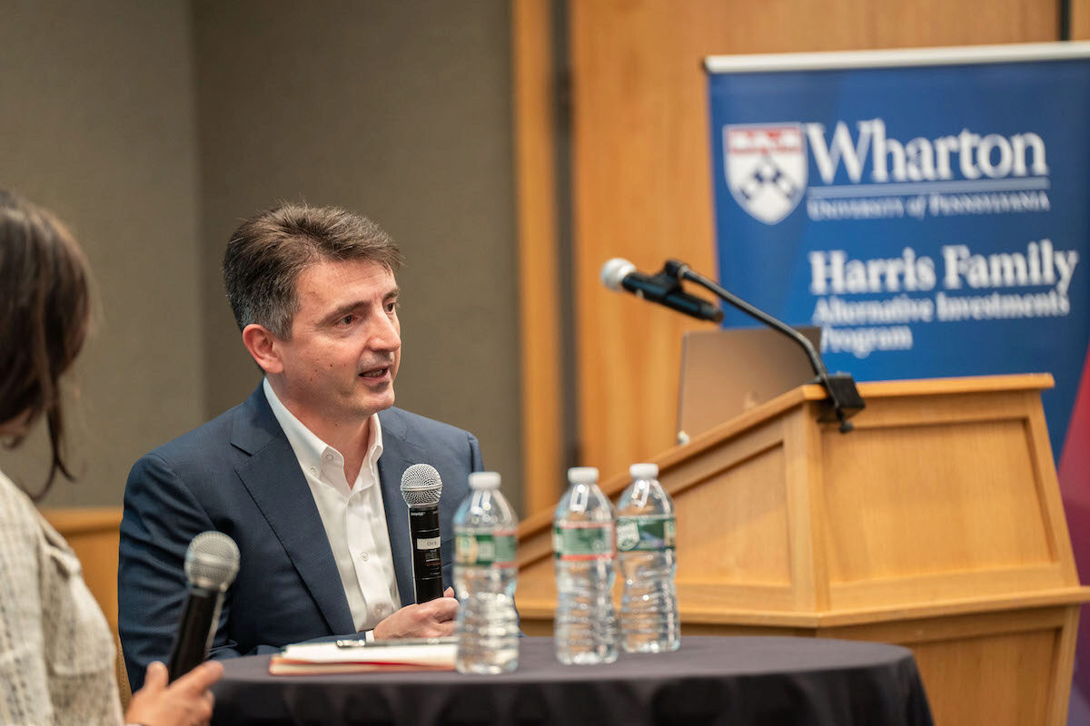 A person in a suit is speaking into a microphone at a panel discussion or seminar. They are seated at a table with several water bottles in front. In the background, there is a banner for the Wharton School's Harris Family Alternative Investment Program.