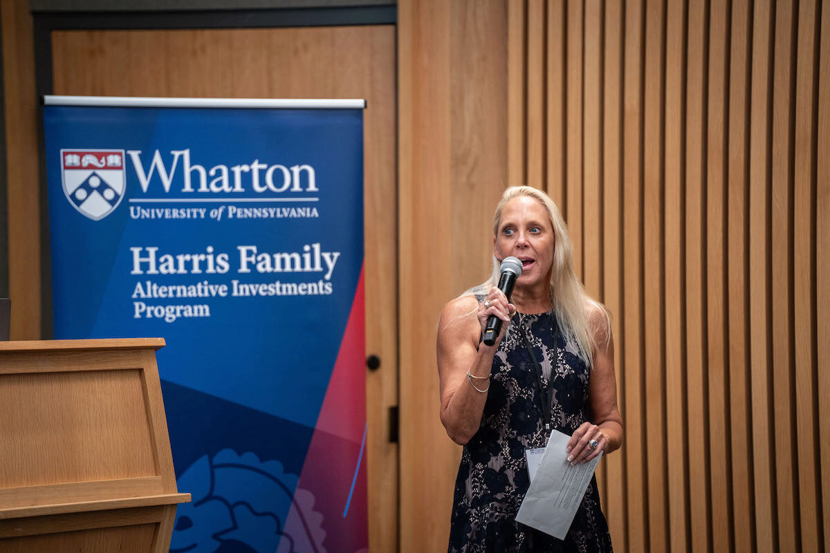 A person speaking into a microphone stands next to a banner for the Wharton University of Pennsylvania Harris Family Alternative Investments Program.