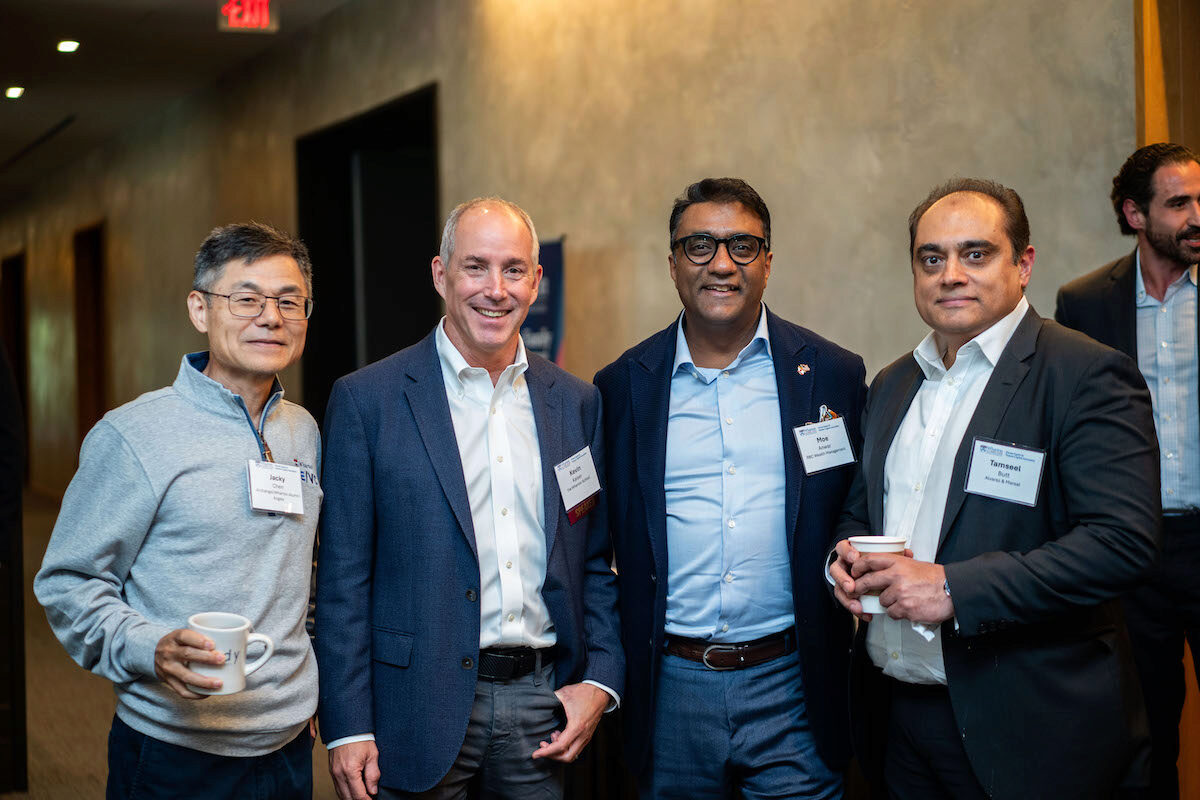 A group of four people in business attire, wearing name tags, standing together indoors. Some are holding coffee cups, suggesting a professional networking event or conference.