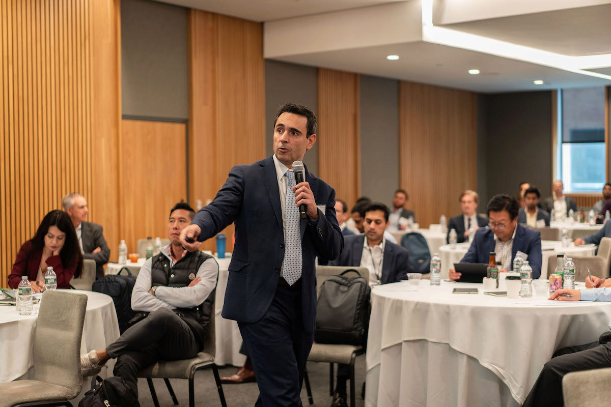 A person in a suit is speaking and gesturing with a microphone in hand at a conference or seminar. Several audience members are seated at round tables with water bottles and notebooks, some appear to be taking notes. The setting is a formal conference room with