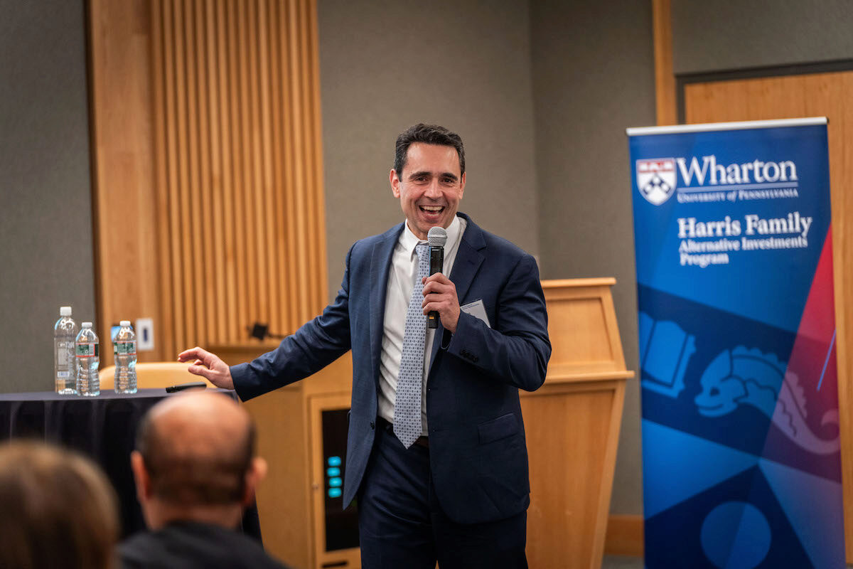 A person in a suit is speaking into a microphone at a professional event, standing beside a Wharton School banner that reads "Harris Family Alternative Investments Program."