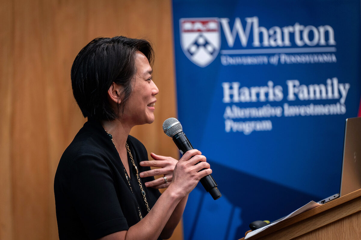 A person speaking into a microphone at a podium, with a background banner displaying "Wharton University of Pennsylvania Harris Family Alternative Investments Program."