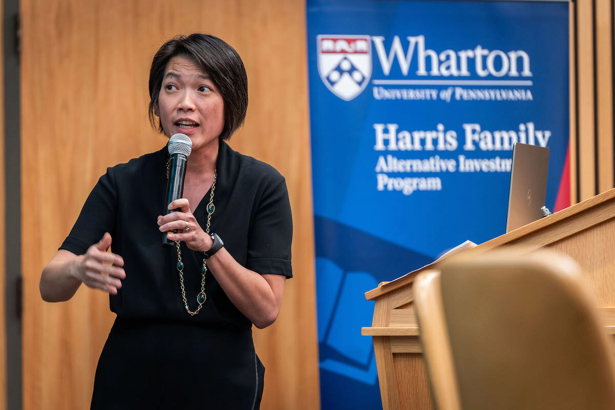 A person is speaking into a microphone, standing next to a podium, with a Wharton School banner in the background.