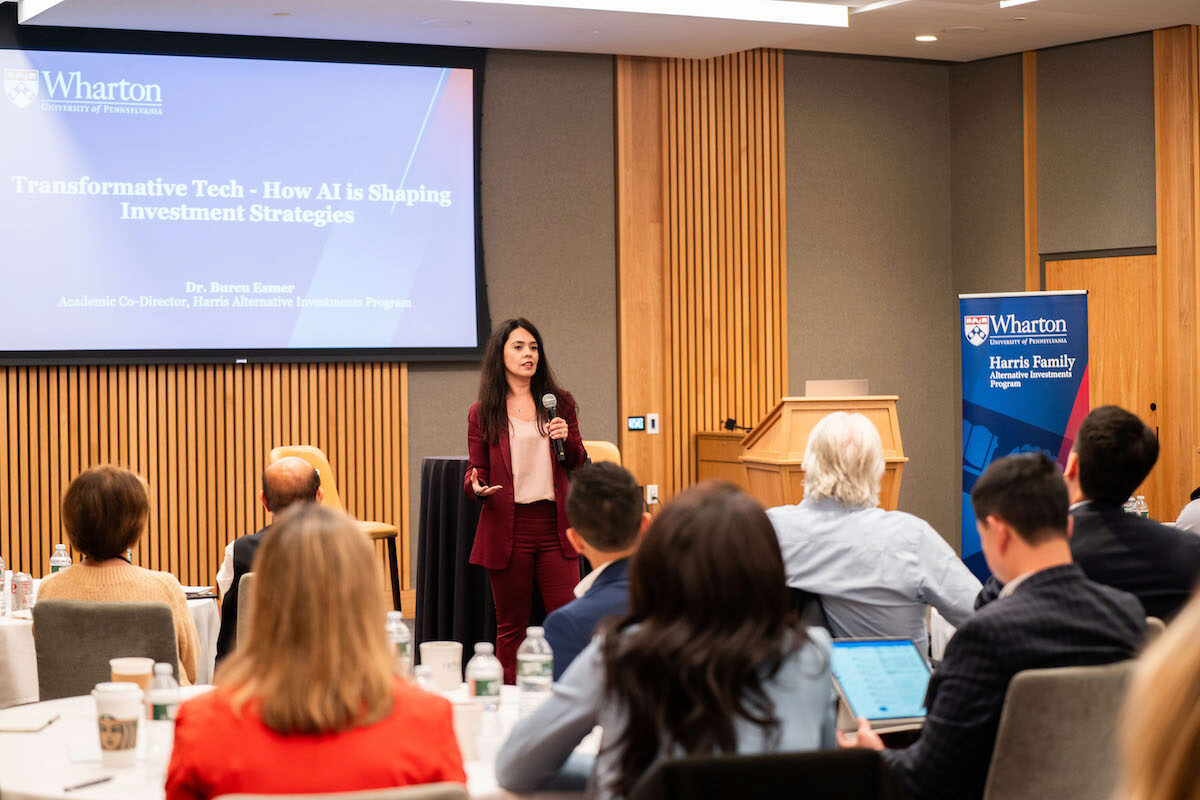 A speaker is presenting to an audience in a conference room. The presentation slide reads "Transformative Tech - How AI is Shaping Investment Strategies." The setting includes a Wharton School banner and individuals seated at tables.