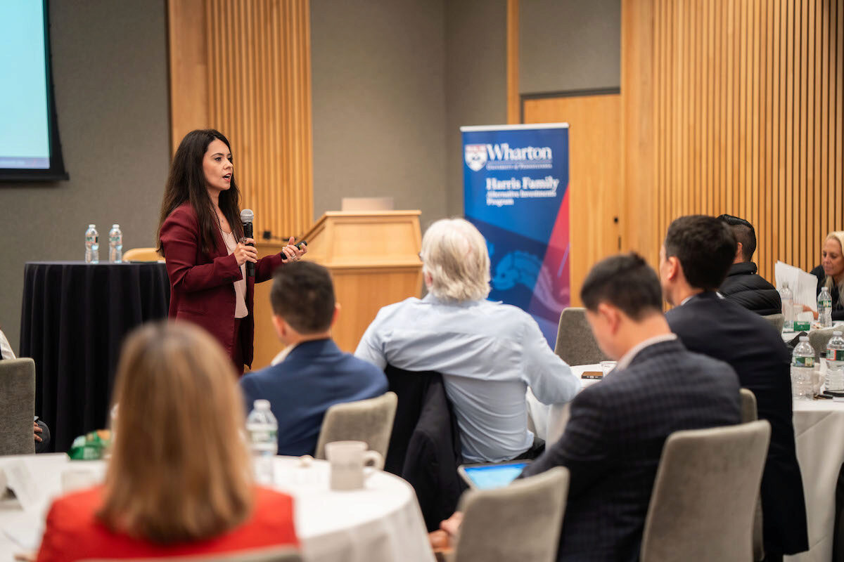 A person is giving a presentation at a conference, speaking into a microphone. Attendees are seated around tables, listening attentively. The setting is professional, with a banner in the background indicating affiliations.