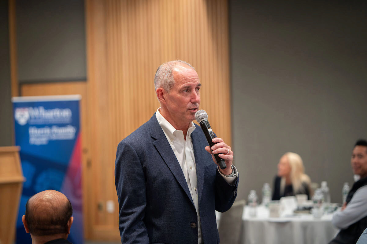 A person in a suit is speaking into a microphone in a conference or meeting setting. In the background, there are attendees seated at tables, and a banner is partially visible.