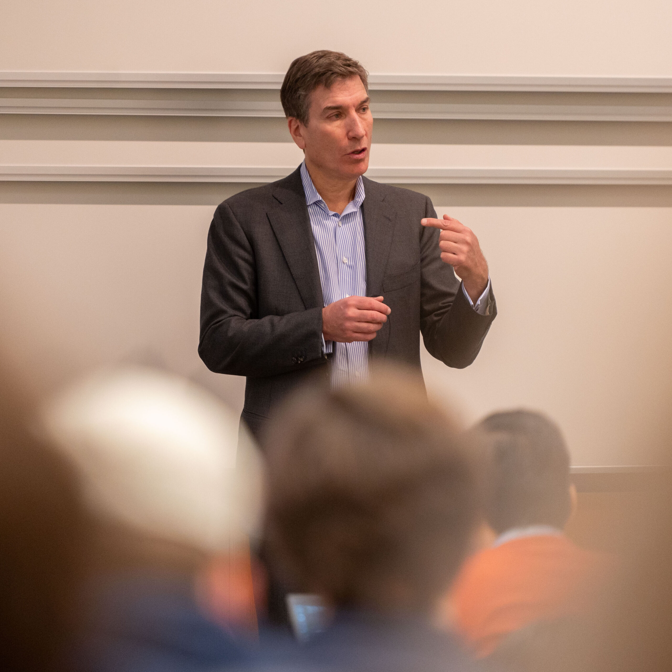 A person in a suit is giving a presentation or lecture in front of an audience. They are gesturing while speaking. The background is a plain wall.