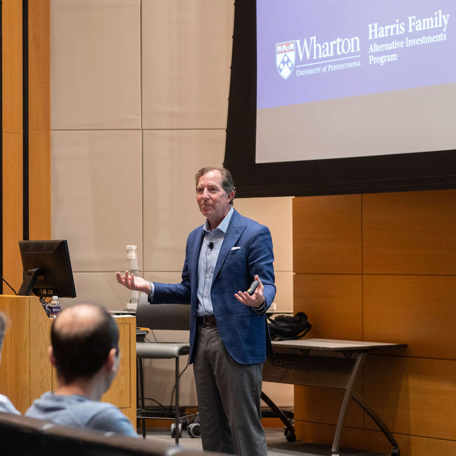 A person speaks to an audience, while gesturing with his hands into a lapel microphone in an academic setting. In the background is a blue screen with the Harris Family Alternative Investments Program logo