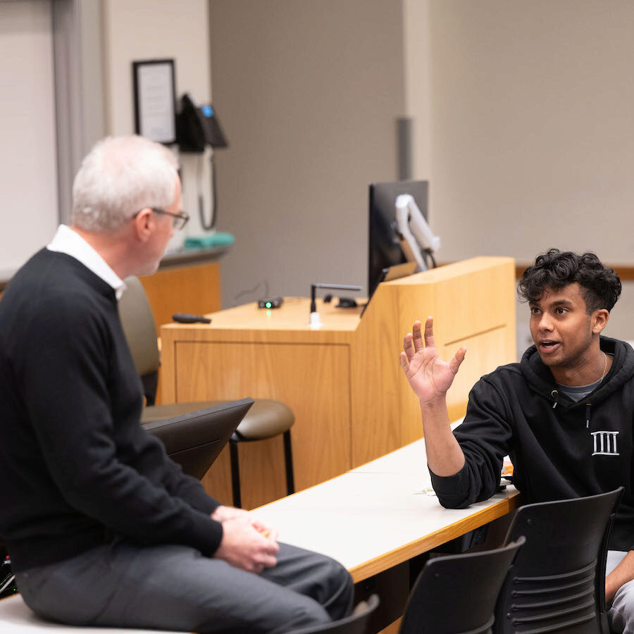 A person sitting on a classroom desk in mid conversation with a person sitting at the desk. The speaker has one hand raised.