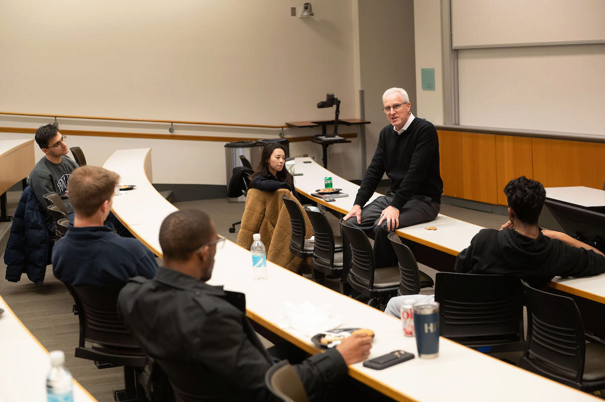 A person sits on a classroom table, addressing a room full of students