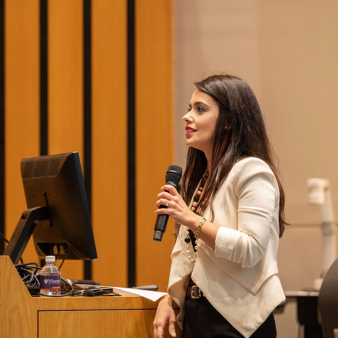 A person in a blazer is speaking into a microphone at a podium with a computer monitor