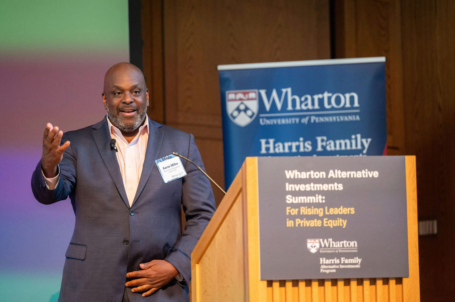 A person speaking at a podium during the Wharton Alternative Investments Summit. The background features a Wharton banner.